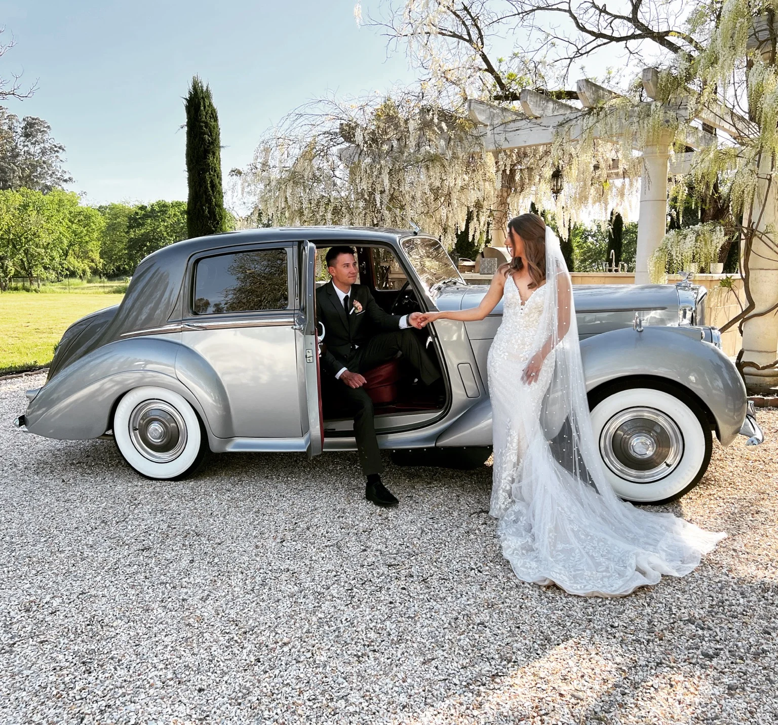 A couple holding hands beside the open Bentley door in a flowering garden