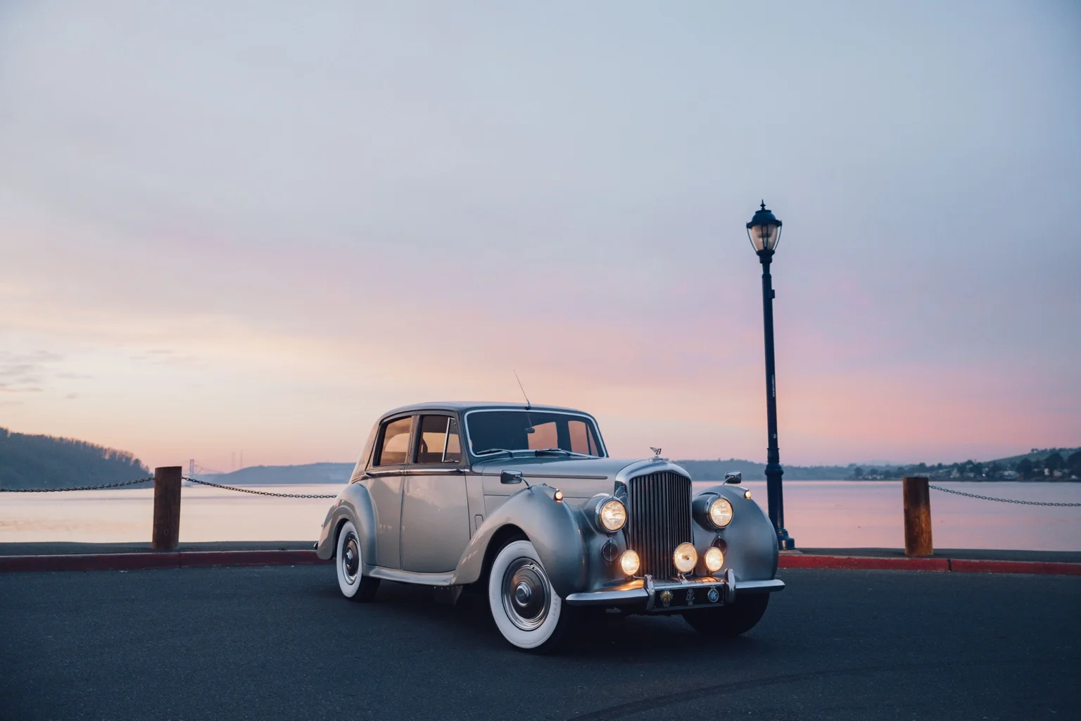 The platinum 1954 Bentley at sunset beside the water, its headlights glowing